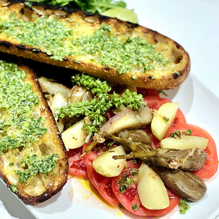 Grilled baguette slice with a touch of pesto, served alongside tomato slices and garlic confit